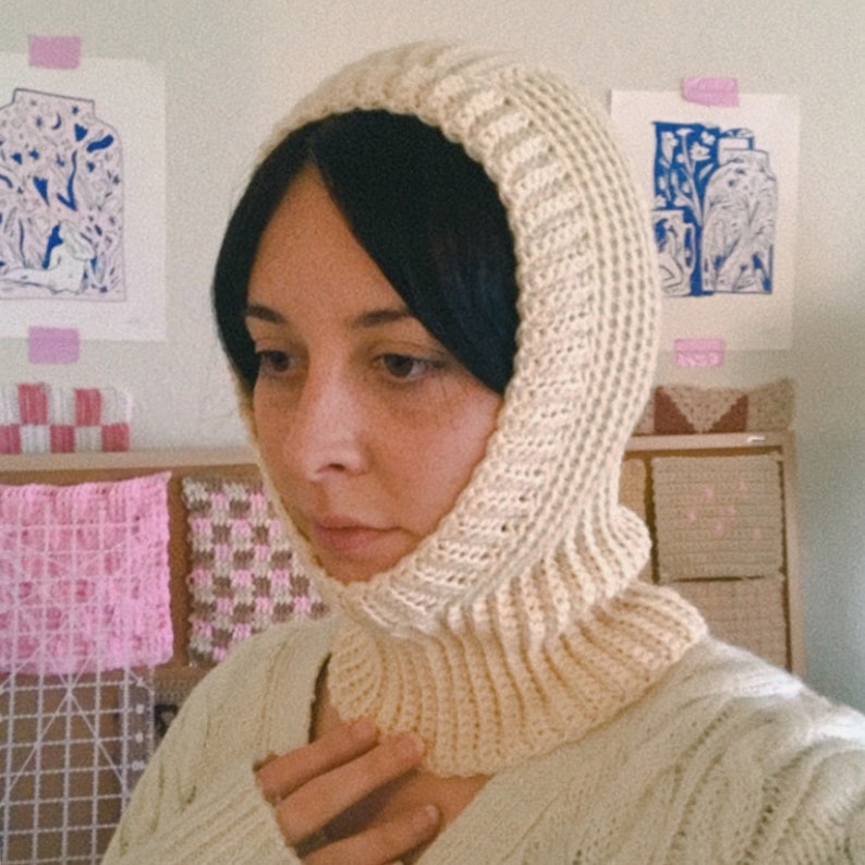 May include: A close-up of a person wearing a white crocheted balaclava. The balaclava is a hooded neck warmer with a ribbed texture. The person is wearing a white sweater.