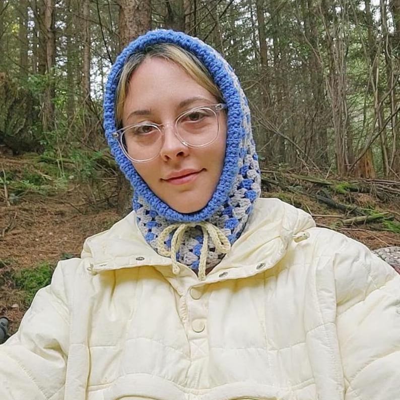 Op de afbeelding: Een persoon draagt ​​een blauwe en witte gehaakte muts met een gele stropdas. De muts is over het hoofd getrokken en bedekt de oren. De persoon draagt ​​een wit jack.