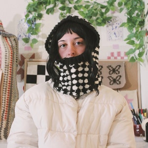 Op de afbeelding: Een persoon draagt een witte pufferjas met een zwarte en witte gebreide cowl-hals. De cowl-hals is een dikke, grove brei met een geometrisch patroon.