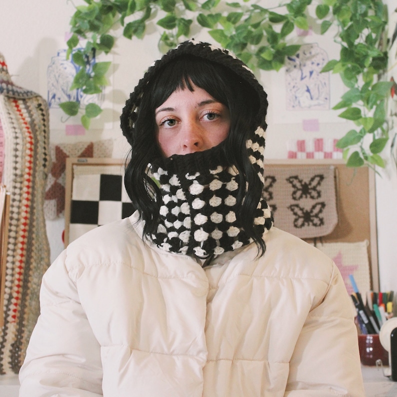 Op de afbeelding: Een persoon draagt een witte pufferjas met een zwarte en witte gebreide cowl-hals. De cowl-hals is een dikke, grove brei met een geometrisch patroon.