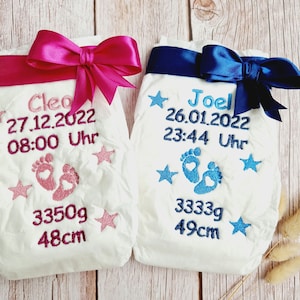 Könnte beinhalten: Zwei weiße Babywindeln mit rosa und blauen Bändern, bestickt mit Namen, Geburtsdatum, Uhrzeit, Gewicht und Größe. Die Windel links zeigt "Cleo 27.12.2022 08:00 Uhr 3350g 48cm" und die Windel rechts zeigt "Joel 26.01.2022 23:44 Uhr 3333g 49cm".
