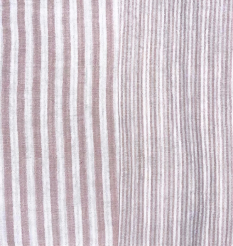 May include: A textile fabric featuring a vertical striped pattern in muted mauve and white. The left side displays wider, soft-edged stripes, while the right side transitions into a denser, finer arrangement of the same mauve and white vertical stripes.