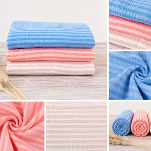May include: A collection of soft, crinkled muslin fabric pieces featuring horizontal stripes in shades of blue, coral, and light pink. The fabric is shown both folded in neat stacks and rolled into cylinders, displaying a lightweight, textured weave.