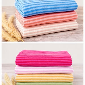May include: A collection of seven folded fabric pieces stacked in two groups. Each piece features a horizontal striped pattern in various colors including blue, orange, light pink, bright pink, pale pink, yellow, and light green with a textured finish.