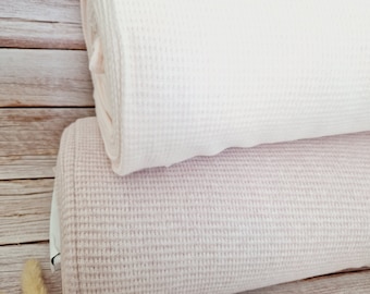 May include: Two rolls of waffle knit fabric stacked horizontally. The top roll is a solid off-white colour, while the bottom roll is a soft, muted dusty pink. Both rolls feature a distinct grid-like texture characteristic of waffle weave fabric.
