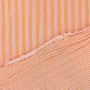 May include: A piece of fabric featuring a pattern of vertical stripes in alternating shades of light orange and soft peach. The material has a visible texture and a frayed, raw edge that runs diagonally across the center of the frame.