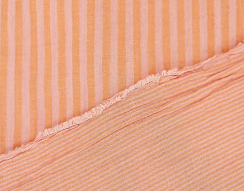 May include: A piece of fabric featuring a pattern of vertical stripes in alternating shades of light orange and soft peach. The material has a visible texture and a frayed, raw edge that runs diagonally across the center of the frame.