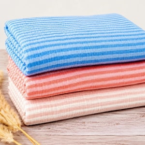 May include: A stack of three folded pieces of fabric featuring a horizontal striped pattern. The top piece is blue and light blue, the middle piece is coral and light pink, and the bottom piece is light pink and white. The fabric has a textured surface.