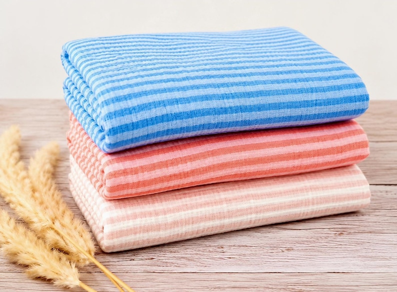 May include: A stack of three folded pieces of fabric featuring a horizontal striped pattern. The top piece is blue and light blue, the middle piece is coral and light pink, and the bottom piece is light pink and white. The fabric has a textured surface.