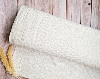 May include: A roll of off-white fabric featuring a subtle, repeating light beige speckled pattern. The textile has a distinct crinkled, textured surface with vertical lines running throughout the material, creating a soft and layered appearance.