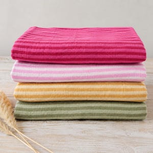 May include: A stack of four folded rectangular textiles featuring horizontal stripes. The top fabric is dark pink, followed by light pink, mustard yellow, and sage green. Each piece has a textured, crinkled appearance with consistent striping.