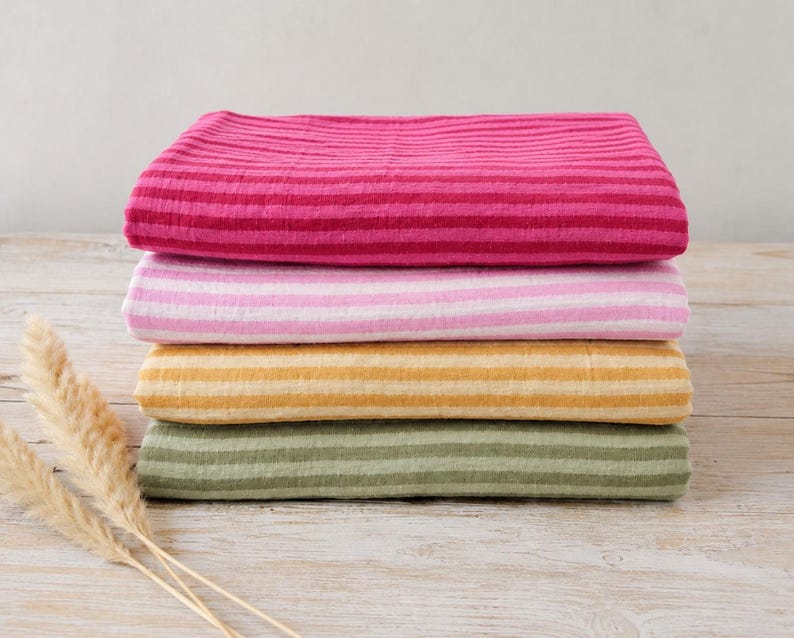 May include: A stack of four folded rectangular textiles featuring horizontal stripes. The top fabric is dark pink, followed by light pink, mustard yellow, and sage green. Each piece has a textured, crinkled appearance with consistent striping.