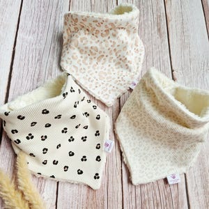 Könnte beinhalten: Drei Baby-Lätzchen mit Cordstruktur und weichem, flauschigem Futter. Die Lätzchen haben eine cremefarbene Farbe mit Leopardenmustern in Braun- und Schwarztönen. Die Lätzchen sind auf einer Holzoberfläche angeordnet.