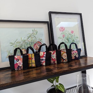 May include: A set of seven small, black fabric tote bags with colourful Japanese fabric panels. The bags are lined up on a wooden shelf with a painting in a frame behind them.