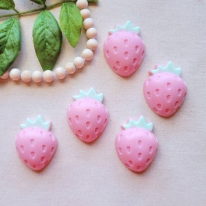 May include: Five pink and white strawberry shaped resin embellishments. The strawberries have a light green top and a pink body with white seeds.