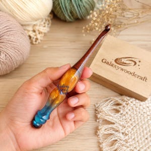 May include: A handcrafted crochet hook featuring a dark brown wooden shaft and a contoured handle made of swirling amber and blue resin. The handle is imprinted with the text Galaxy Wood Craft. The hook is designed for yarn work and fiber arts.