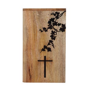 May include: A wooden plaque with a black silhouette of a cross and a floral branch design. The plaque has a natural wood grain finish.