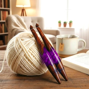 May include: Two wooden crochet hooks with purple resin detailing are placed near a ball of cream-coloured yarn. The hooks feature a dark wood grain and a decorative purple section. A mug of tea and a book are in the background, suggesting a cosy crafting scene.