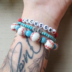 May include: A layered bracelet set with white beads that resemble baseballs, blue turquoise beads, and white letter beads spelling "JACKSON IS". The bracelet is on a person's wrist with a tattoo.