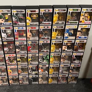 Puede incluir: Una colección de figuras de vinilo Funko Pop! en sus cajas originales. Las figuras representan personajes de varias franquicias, incluyendo Marvel y DC Comics. Las cajas negras tienen ventanas transparentes.