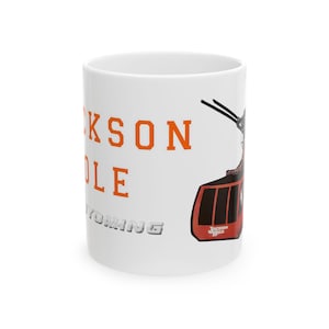May include: A white ceramic mug featuring the orange text JACKSON HOLE and the grey text WYOMING. To the right, a graphic illustration of a red aerial tram car is depicted with black cables extending upwards, including a small logo on the tram.