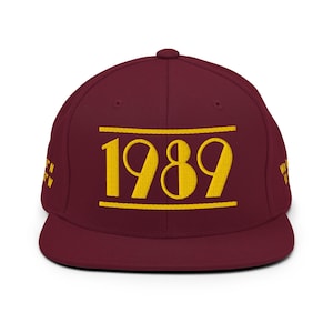Puede incluir: Una gorra snapback color granate con el texto amarillo bordado 1989 centrado entre dos líneas horizontales. Los lados de la gorra muestran texto amarillo bordado adicional, incluyendo coordenadas a la izquierda y texto parcial a la derecha.