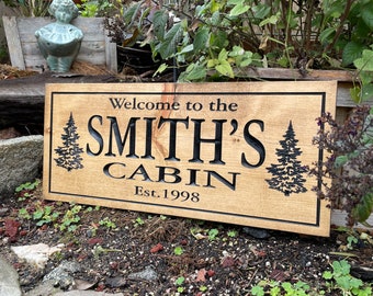 Puede incluir: Un letrero de madera con el texto "Welcome to the Smith's Cabin Est. 1998" en letras negras. El letrero tiene dos siluetas de pinos a cada lado del texto.