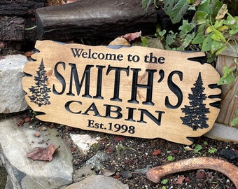 Puede incluir: Un letrero de madera rústica con la inscripción "Welcome to the Smith's Cabin Est. 1998" en letras negras. El letrero presenta dos pinos estilizados a cada lado del texto.