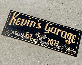 Puede incluir: Un letrero de madera negro con el texto "Kevin's Garage Est. 2023" en letras doradas. El letrero tiene un diseño de llamas y llaves cruzadas.