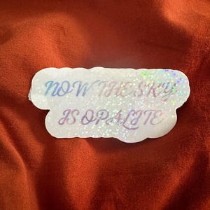 May include: A white sticker with the text "NOW THE SKY IS OPALITE" in a holographic, iridescent font. The sticker has a cloud-like shape and is set against a reddish-brown background.