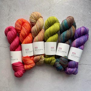 Könnte beinhalten: Eine Sammlung bunter Garnknäuel in Pink, Orange, Gelb, Grün, Blau und Lila. Jedes Knäuel hat ein weißes Etikett mit dem Text "HOT MAGENTA YARN COMPANY". Das Garn liegt auf einer hellgrauen Oberfläche.