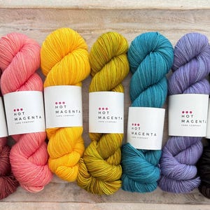 Puede incluir: Una colección de ovillos de lana coloridos en tonos rosa, amarillo, verde, turquesa y morado. Cada ovillo tiene una etiqueta blanca con el texto "HOT MAGENTA YARN COMPANY". La lana está dispuesta sobre una superficie de madera clara.