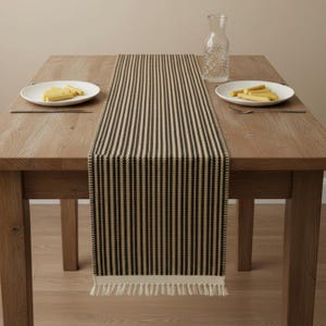May include: A black and beige striped table runner with a fringed edge is displayed on a wooden table. Two white plates with food and a clear glass water carafe are also on the table. The runner's vertical stripes create a simple, modern aesthetic.