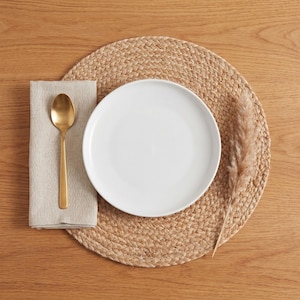 May include: A white ceramic plate, gold spoon, and beige linen napkin rest on a round woven placemat. A dried ornamental grass stem is placed on the right side. The items are arranged on a light brown wooden surface.