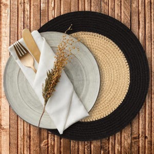 May include: A rustic table setting featuring a light gray plate, a white folded napkin, and gold-colored cutlery. A sprig of dried flowers adds a natural touch. The setting rests on a woven placemat with a black border, set against a wooden background.