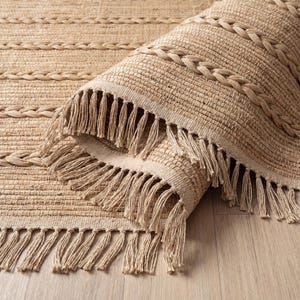 May include: A rolled-up, beige woven rug with braided and fringed edges. The rug features a textured design with horizontal lines and decorative braided accents. The fringe hangs down, adding a decorative element to the rug.