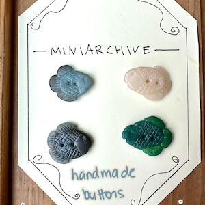 May include: Four handmade fish-shaped buttons in shades of blue, pink, gray, and green, arranged on a white card with the text "MINIARCHIVE" and "handmade buttons (set of 4)". The buttons are displayed on a wooden surface.