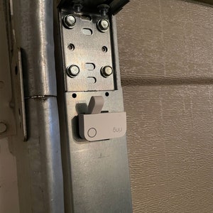 May include: A white garage door opener sensor with a black circle and the word "Ring" printed on it. The sensor is attached to a metal garage door track.