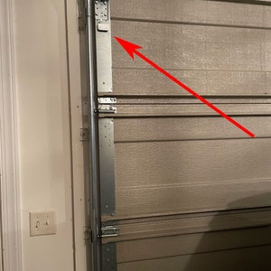 May include: Close-up of a garage door opener mechanism with a metal track and a white sensor. The sensor is attached to the track with screws.
