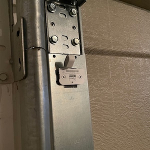 May include: Close-up of a silver metal garage door opener bracket with a white sticker that says "Garage Door Plus" and a black rubber band. The bracket is attached to a brown garage door.
