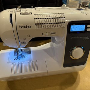 May include: A white Brother sewing machine with a black control panel, a digital display showing 01, and the text STRONG & TOUGH ST150HDH. The machine features a stitch selection chart on the top, a needle area with blue light, and a ruler.