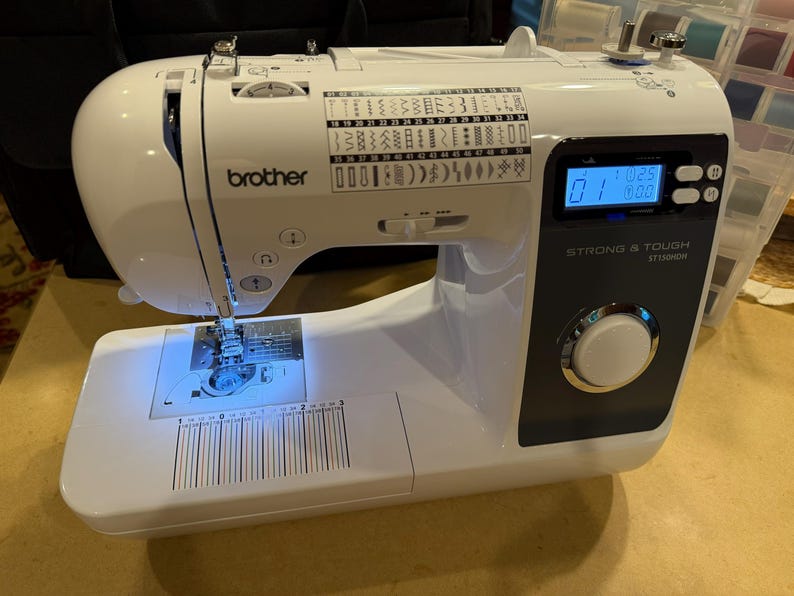 May include: A white Brother sewing machine with a black control panel, a digital display showing 01, and the text STRONG & TOUGH ST150HDH. The machine features a stitch selection chart on the top, a needle area with blue light, and a ruler.