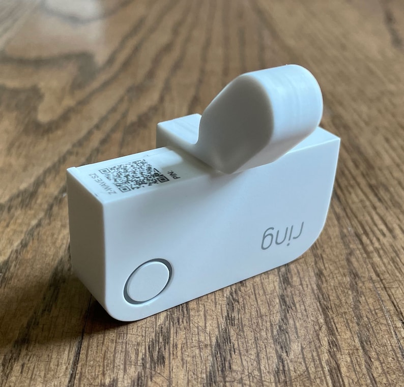May include: White Ring doorbell with a QR code and the word "Ring" printed on the side. The doorbell has a small, round button on the front.