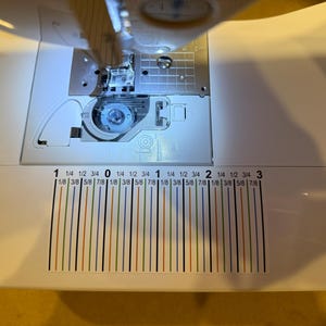 May include: A white sewing machine bed featuring a clear plastic bobbin cover and a printed seam guide decal. The decal displays black numerical markings from 1 to 3 with fractional inch increments indicated by vertical colored lines.