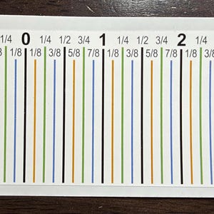 May include: A rectangular white sticker featuring a ruler-style measurement guide with black, green, blue, and orange vertical lines. The text includes numbers 1, 0, 1, 2, 3 and fractions like 1/8, 1/4, 3/8, 1/2, 5/8, 3/4, and 7/8 in black ink.