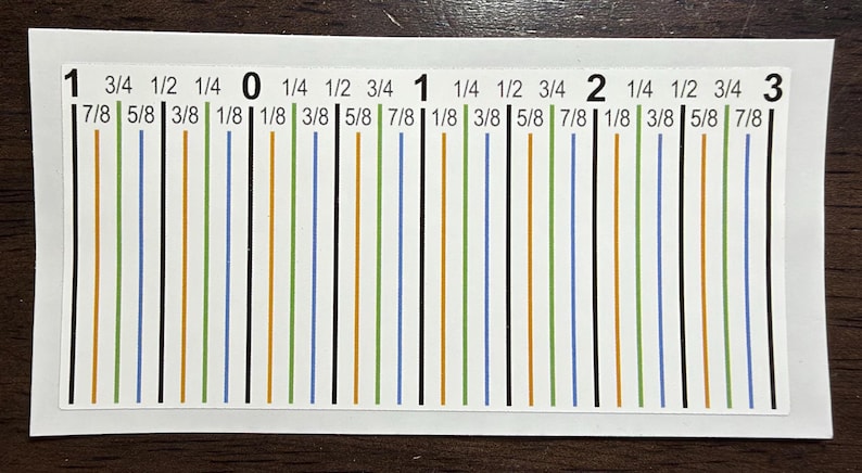 May include: A rectangular white sticker featuring a ruler-style measurement guide with black, green, blue, and orange vertical lines. The text includes numbers 1, 0, 1, 2, 3 and fractions like 1/8, 1/4, 3/8, 1/2, 5/8, 3/4, and 7/8 in black ink.