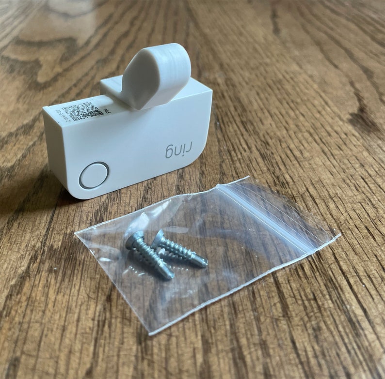 May include: A white Ring doorbell with a QR code and the word "Ring" printed on it. The doorbell is packaged with two small silver screws in a clear plastic bag.