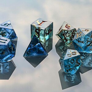 Könnte beinhalten: Ein Satz von sieben Würfeln in verschiedenen Formen, darunter ein d4, d6, d8, d10, d12, d20 und ein Prozentwürfel. Die Würfel sind transparent blau mit goldfarbenen Zahlen und dekorativen Elementen. Die Würfel werden auf einer reflektierenden Oberfläche präsentiert.
