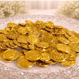 Pode incluir: Um prato de vidro transparente cheio de numerosas moedas douradas. As moedas são redondas e parecem ter desenhos em relevo. O prato tem uma borda decorativa e brilhante. O fundo apresenta flores brancas.