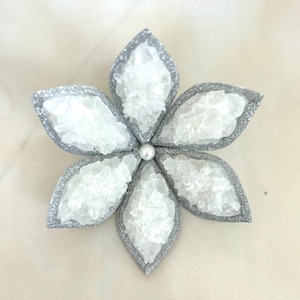 May include: A decorative flower-shaped ornament with five petals. Each petal has a silver glitter border and is filled with white, crystalline material. A small, white pearl is at the center of the flower.